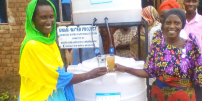 Two women in front of a sawyer filter system in Uganda comparing a glass of clean water with dirty water.